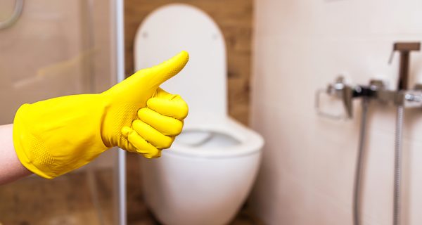 Against the background of a toilet bowl and a bathroom in a modern style, close-up of a hand in a yellow protective glove shows the sign excellent. The concept of good cleaning, indoor hygiene.